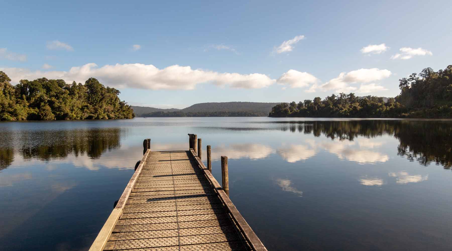 Wooden jetty on still water reflecting the fluffy white clouds on Lake Mapourika West Coast New Zealand