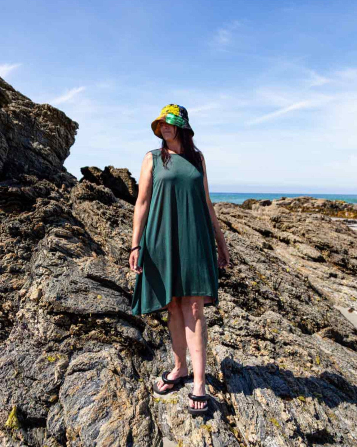 Woman with a multicoloured bucket hat standing on a rocks at a beach wearing the Asmuss Drift Dress without the waist tie that shows how the fabric falls