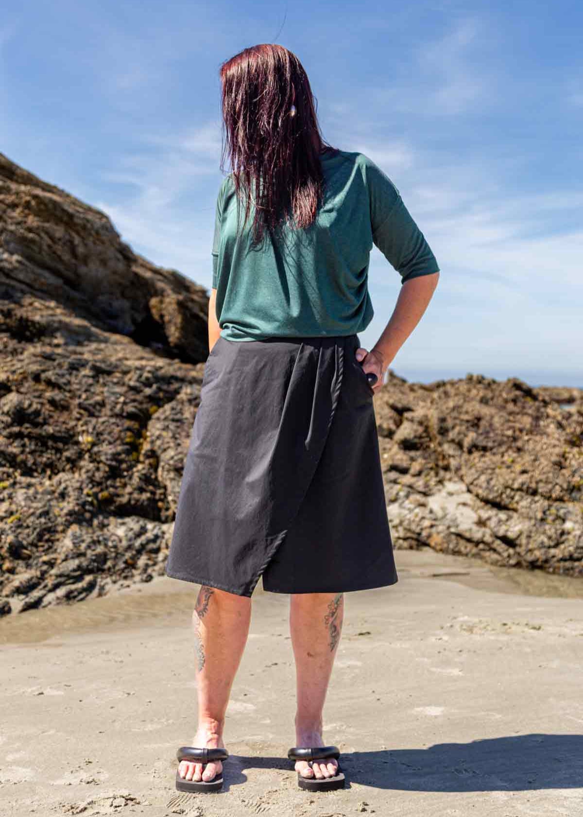 Woman standing in front of big rocks on a beach wearing the black wearing the Evie Wrap Skirt and Vivienne Cowl Neck Top
