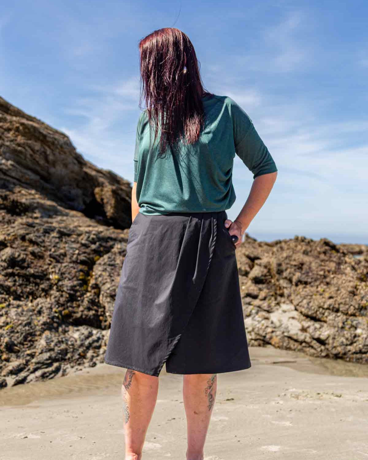 Woman standing  in front of big rocks on a beach wearing the black wearing the Evie Wrap Skirt and Vivienne Cowl Neck Top