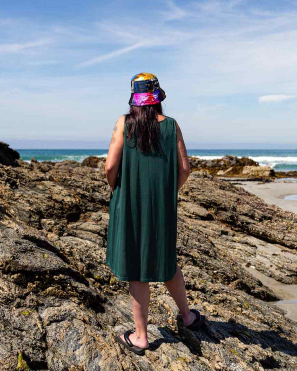 Woman with a multicoloured bucket hat standing on rocks looking out at the ocean wearing the Asmuss Drift Dress without the waist tie that shows how the fabric falls