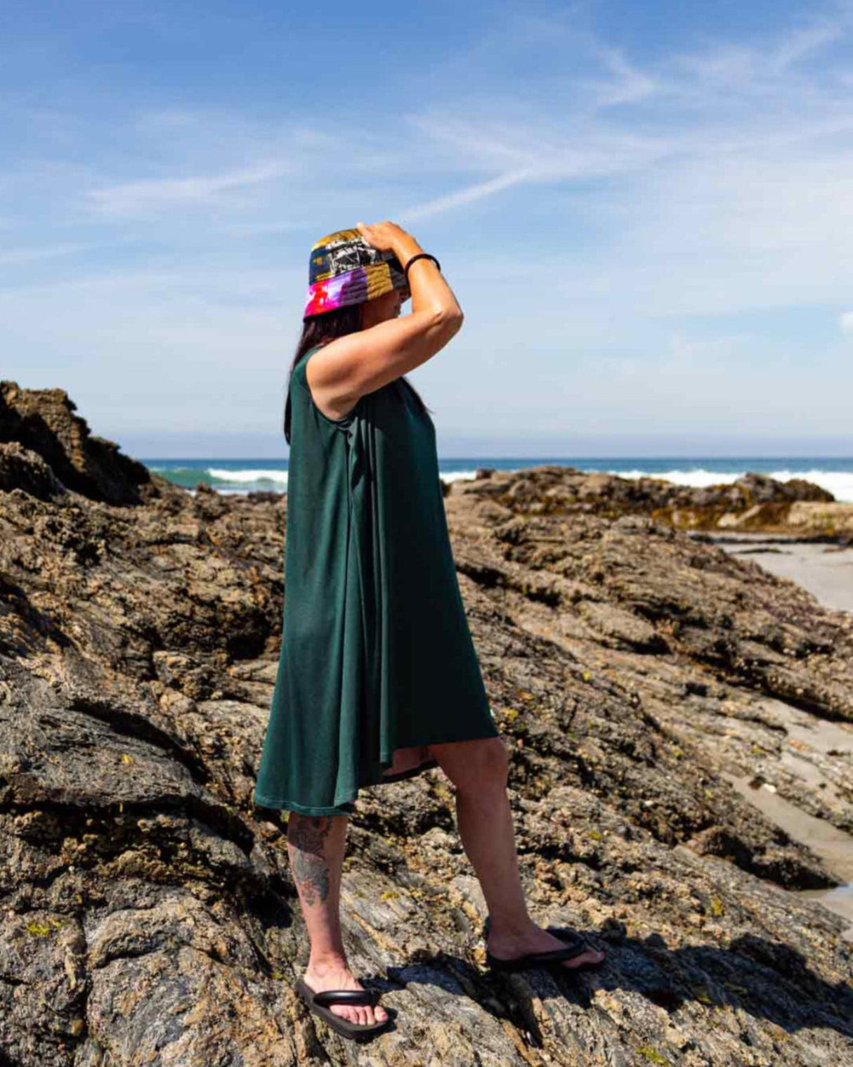 Woman with a multicoloured bucket hat standing on rocks looking out at the ocean wearing the Asmuss Drift Dress show the inverted pleat detail on the side of the dress