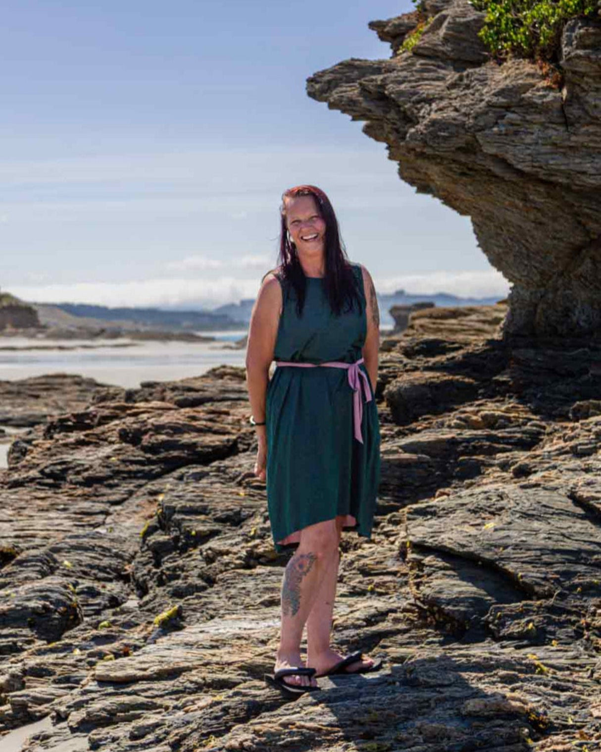 Woman standing on rocks at a beach wearing the Asmuss Drift Dress with the pink waist tie cinching in the dress