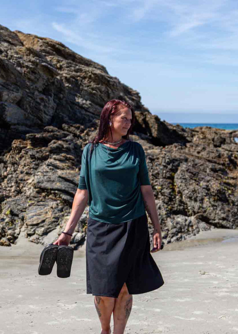 Woman walking on a sandy beach in front of big rocks carrying her flip flops in her hand wearing the black Evie Wrap Skirt and Vivienne Cowl Neck Top