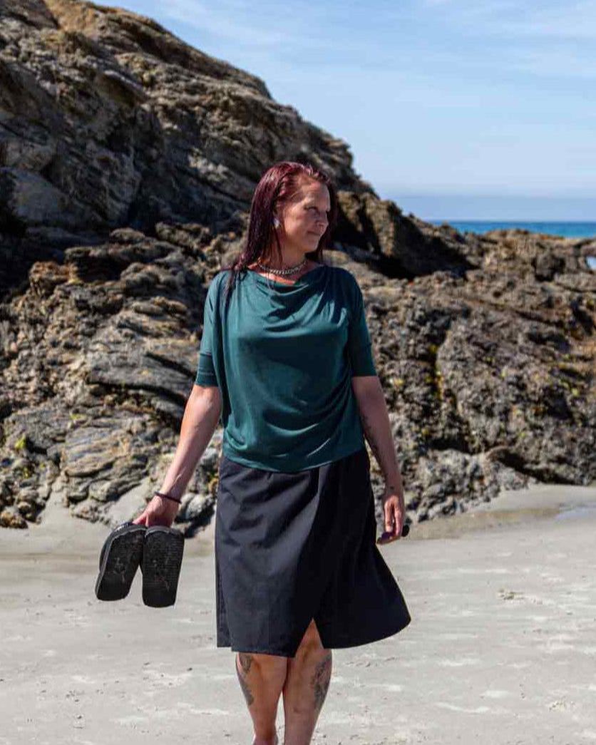 Woman walking on a  sandy beach in front of big rocks carrying her flip flops in her hand wearing the black Evie Wrap Skirt and Vivienne Cowl Neck Top