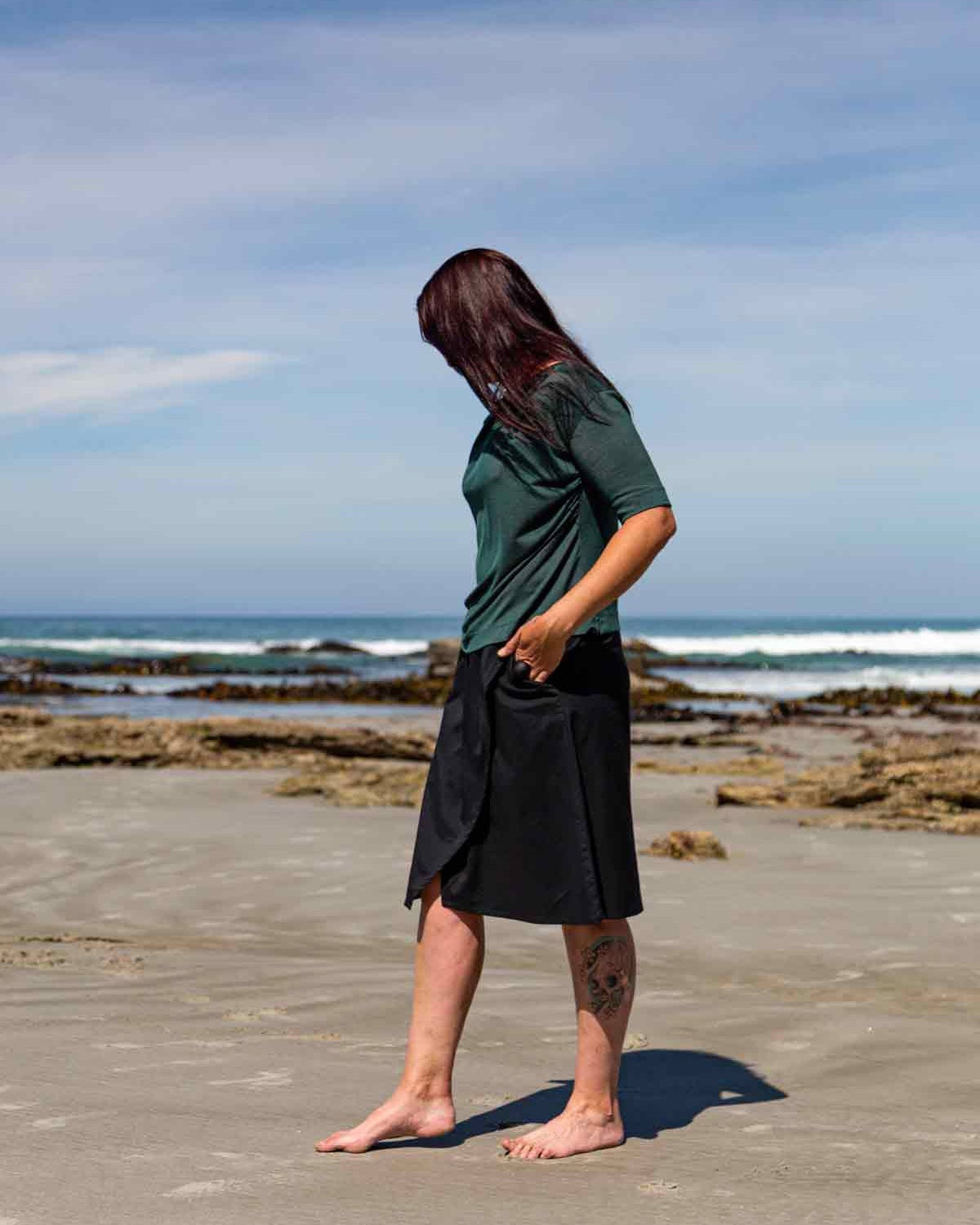Woman walking on a  sandy beach in front of big rocks wearing the black wearing the Evie Wrap Skirt and Vivienne Cowl Neck Top