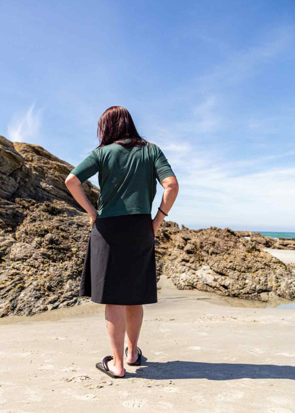 Woman standing on a sandy beach facing away looking at big rocks wearing the black wearing the Evie Wrap Skirt and Vivienne Cowl Neck Top