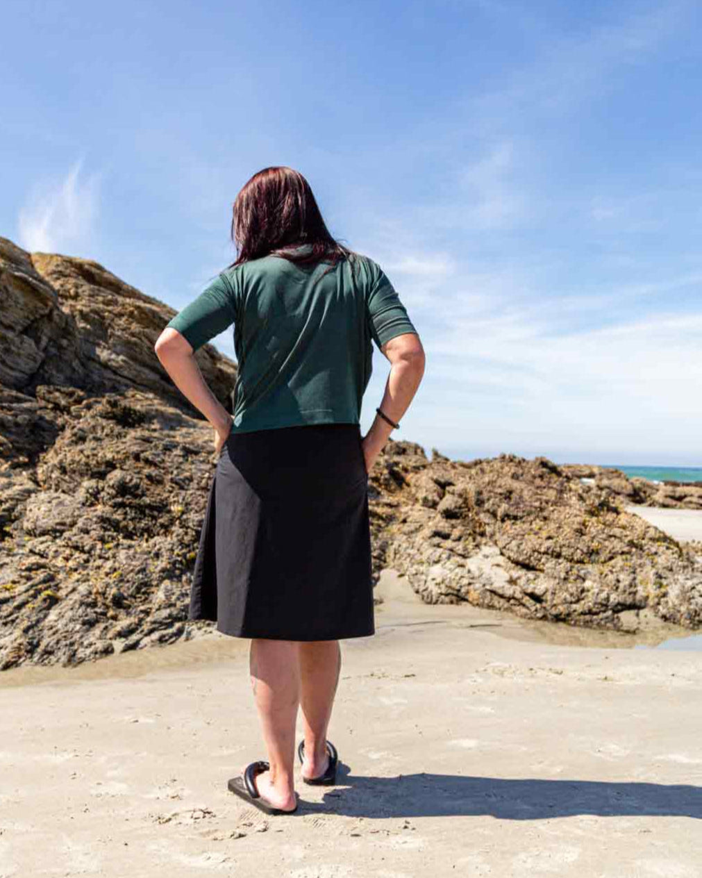 Woman standing on a  sandy beach facing away looking at big rocks wearing the black wearing the Evie Wrap Skirt and Vivienne Cowl Neck Top