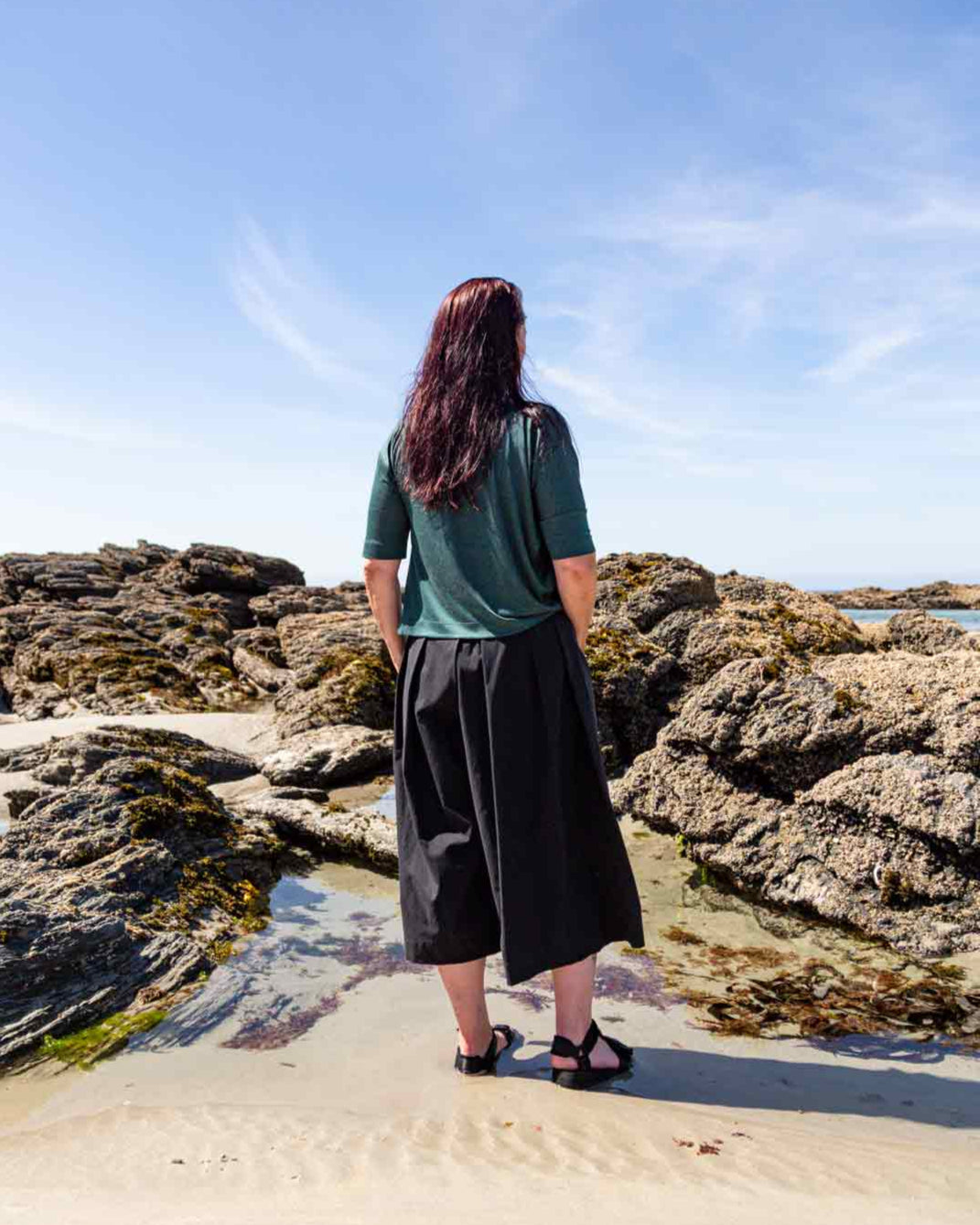 Woman standing in front of rock pools on a sandy beach wearing the black Kate Culottes and green Vivienne Cowl Neck Top showing the pleat details on the back of the culottes