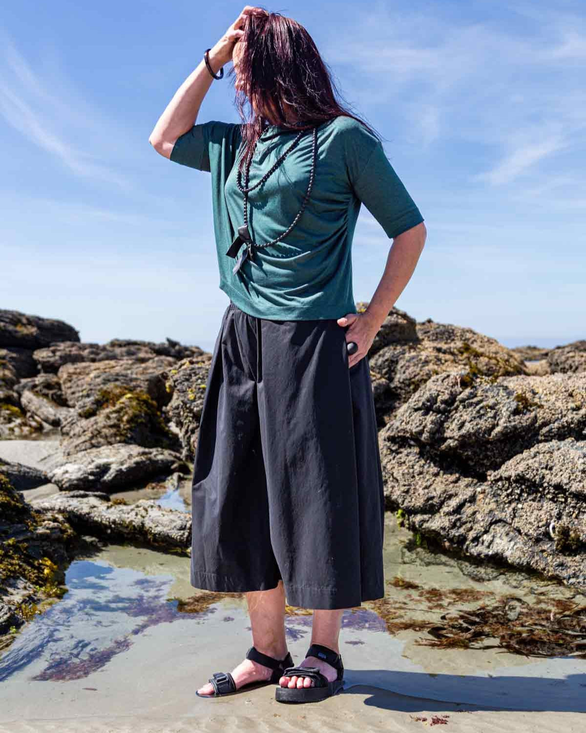 Woman standing in front of rock pools on a sandy beach, with her hand in her hair, wearing the black Kate Culottes and green Vivienne Cowl Neck Top