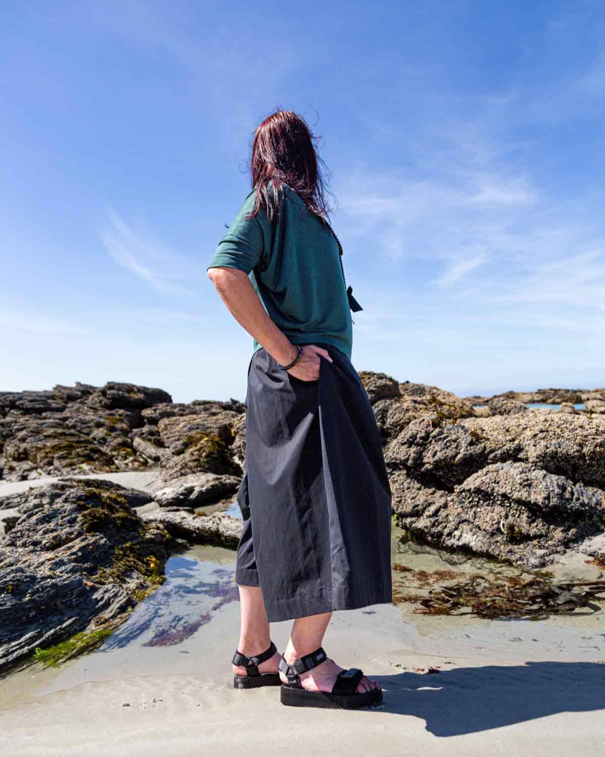 Woman standing side on in front of rock pools on a sandy beach wearing the black Kate Culottes and green Vivienne Cowl Neck Top. She has her hand in one of the pocket sand shows the side pleat in the culottes