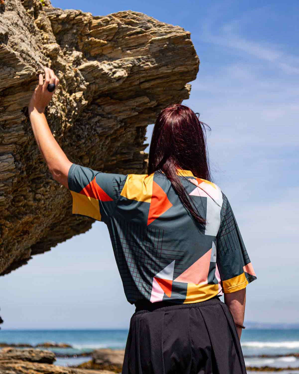 Woman standing looking toward the blue sea with her left hand leaning on rocks the are overhead. She is wearing the Asmuss Drew T-shirt