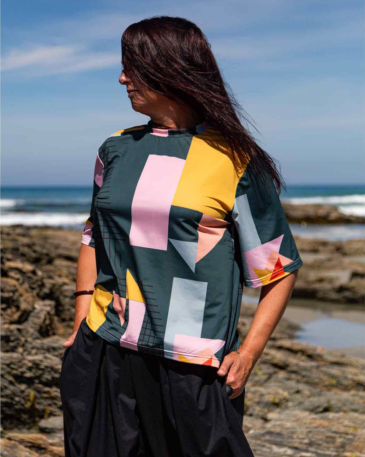 Woman wearing the Asmuss Drew T-shirt. She has long dark hair, is looking right and is standing on rocks in front of the blue sea and blue sky