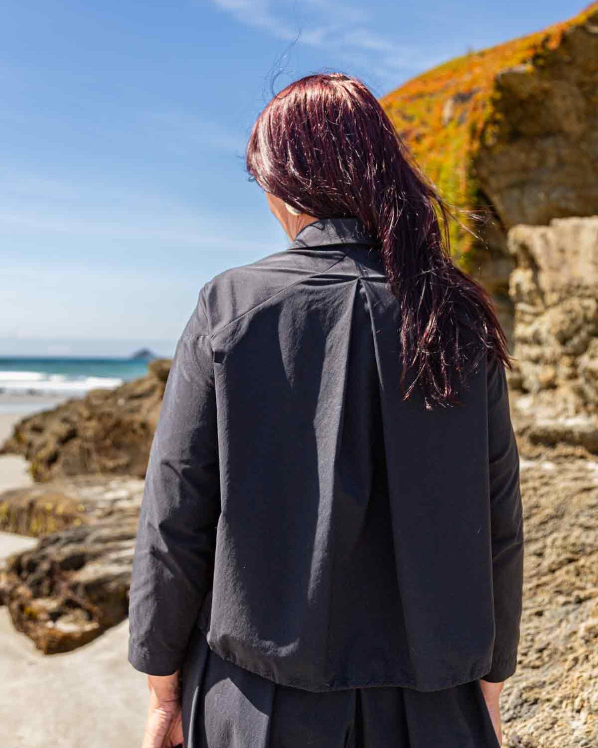 Woman facing towards the sea at a beach with rock outcrops. Her long dark hair is over her right shoulder. She is wearing the Asmuss Roam Jacket with an inverted pleat from the centre back giving it an A line shape