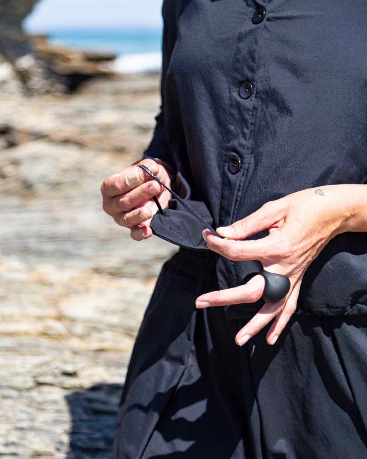 Woman wearing the black Asmuss Roam Jacket showing the drawstring that can be used to create shape in the jacket. The Roam Jacket has a collar, button front and drawstring