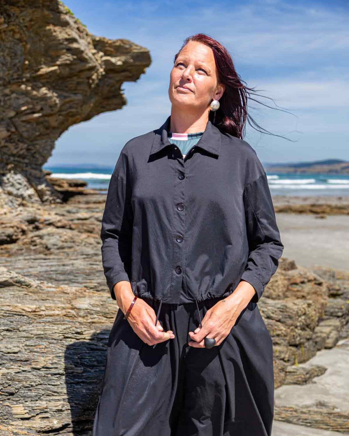 Woman wearing the black Asmuss Roam Jacket showing the drawstring that can be used to create shape in the jacket. The Roam Jacket has a collar, button front and drawstring