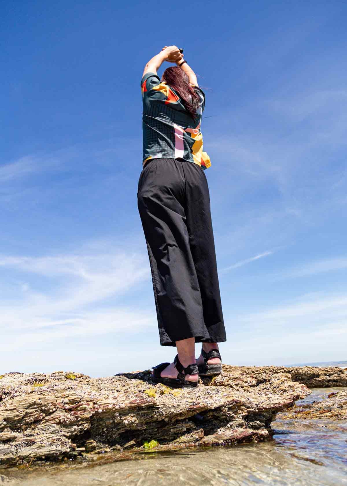 Woman with long dark hair standing on rocks at a beach with her hands head above her head. The photo is taken from below looking up at the black Asmuss Elsie Wide Leg Trousers. She is also wearing the Asmuss Drew T-shirt in a geometric print