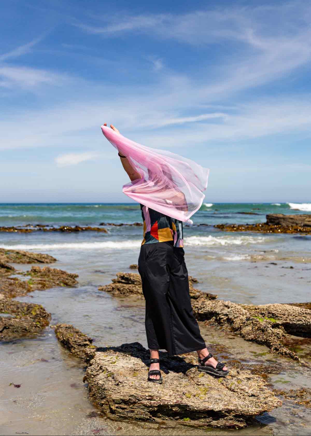 Woman with long dark hair standing on rocks at a beach holding pink tulle in front of her. She is wearing black Asmuss Elsie Wide Leg Trousers and Drew T-shirt in a geometric print