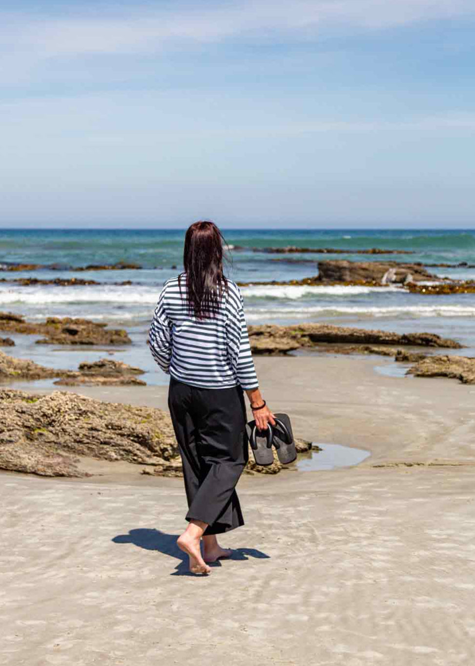 Woman, with long dark hair, walking on the beach towards the water. She is wearing the black Asmuss Elsie Wide Leg Trousers and white and dark green striped Zaha Puzzle Top
