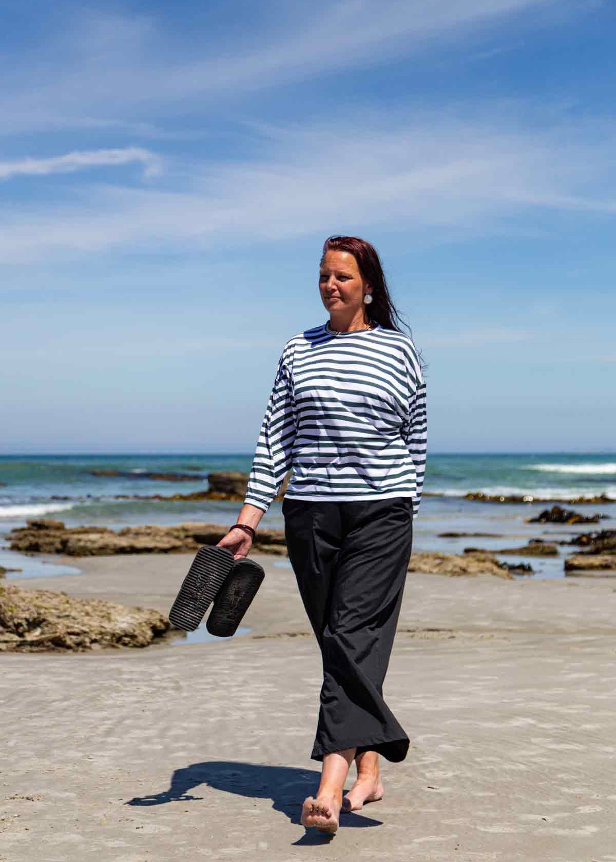 Woman, with long dark hair, walking on a beach with her sandals in her hand. She is wearing the black Asmuss Elsie Wide Leg Trousers and white and dark green striped Zaha Puzzle Top