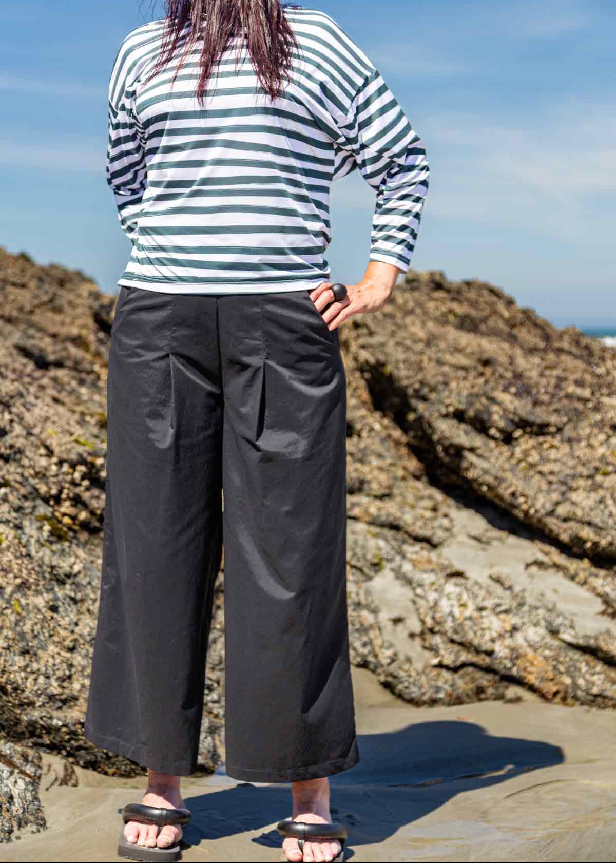 Woman standing in front of rocks on a beach with her hands on her hips. She is wearing the black Asmuss Elsie Wide Leg Trousers and white and dark green striped Zaha Puzzle Top