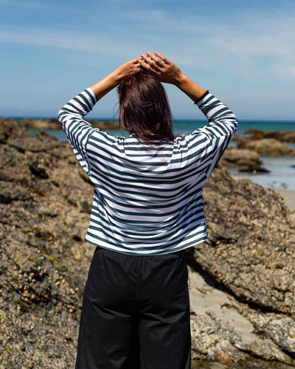 Woman with dark hair facing towards rocks and the blue sea with her hands on her head. She is wearing the Asmuss Zaha Puzzle Top with horizontal painterly stripes in white and dark green.