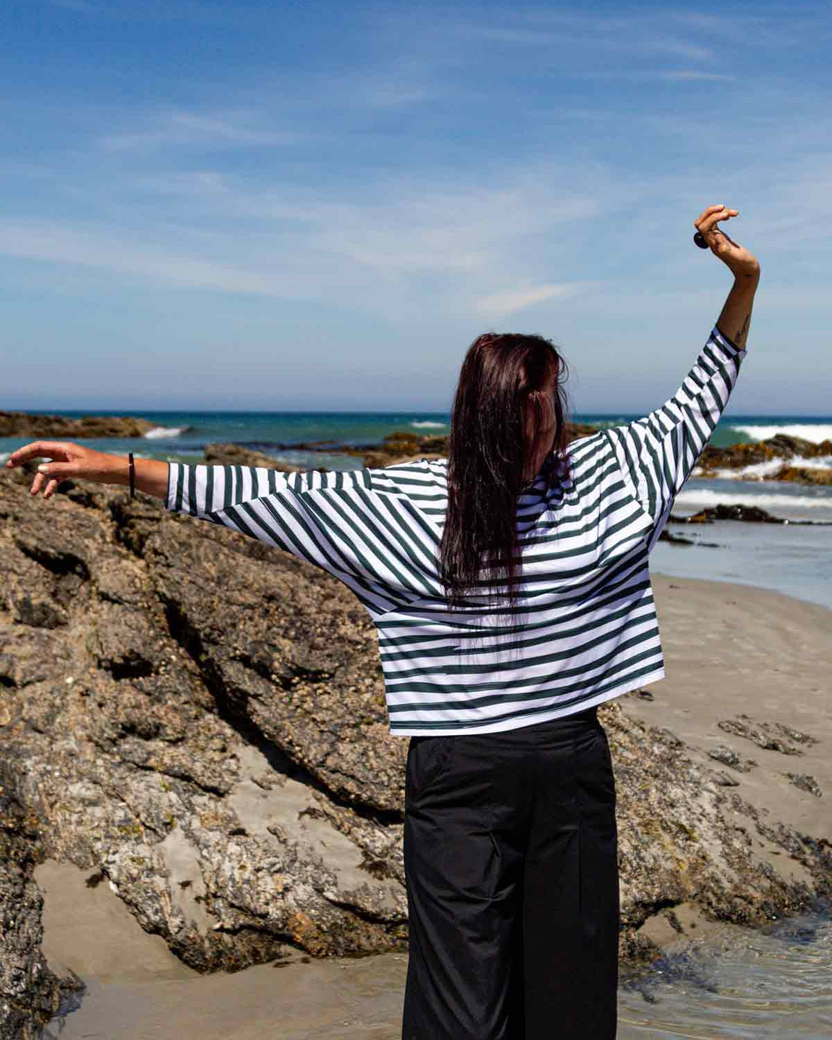 Woman with dark hair standing in front of rocks and the blue sea with her arms held out. She is wearing the Asmuss Zaha Puzzle Top with horizontal painterly stripes in white and dark green