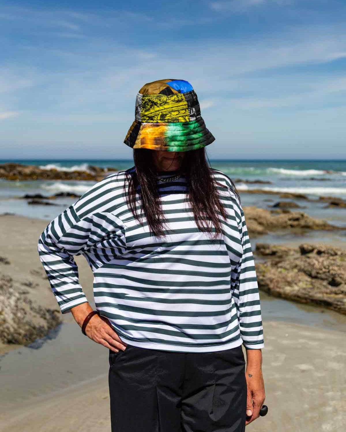 Woman with dark hair standing in front of rocks and the ocean with her right hand on her hip. She is swearing the Asmuss Zaha Puzzle Top with horizontal painterly stripes in white and dark green and a brightly coloured bucket hat