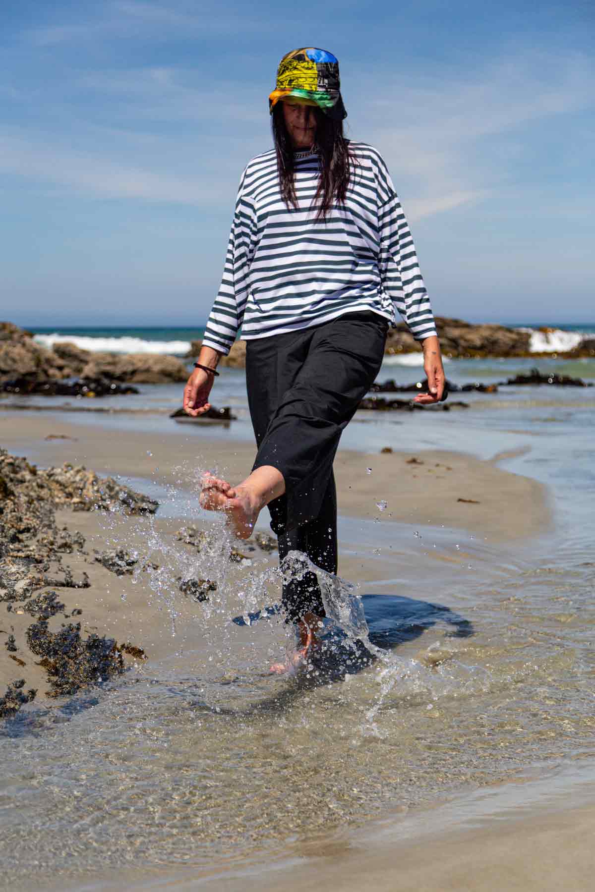 Woman, with long dark hair, kicking up water as she walks through it. She is wearing the black Asmuss Elsie Wide Leg Trousers and white and dark green striped Zaha Puzzle Top