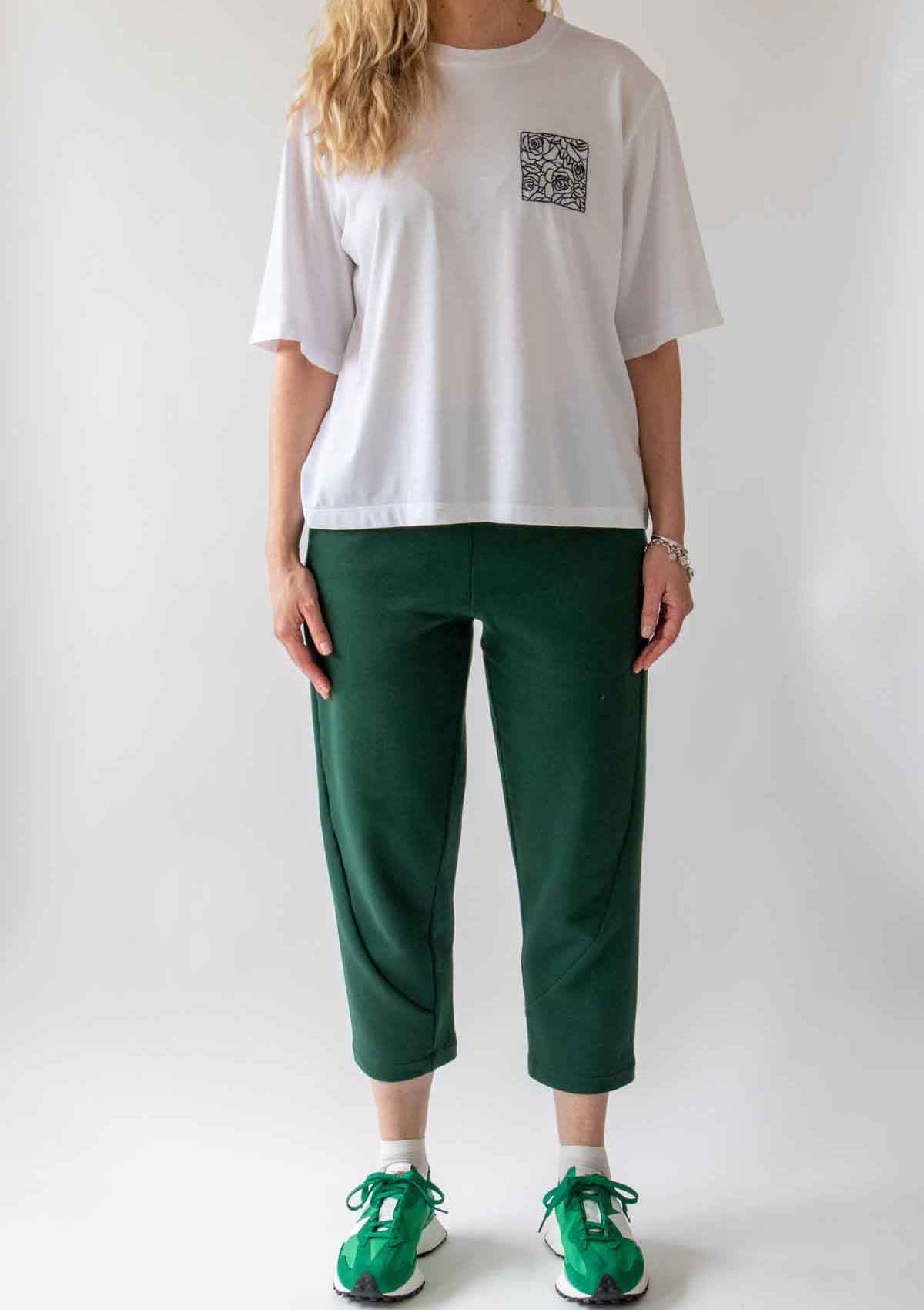 Woman standing front on wearing the Asmuss Curve Joggers in Trekking Green and Evelyn Tshirt in White with Black Rose Embroidery and bright green trainers