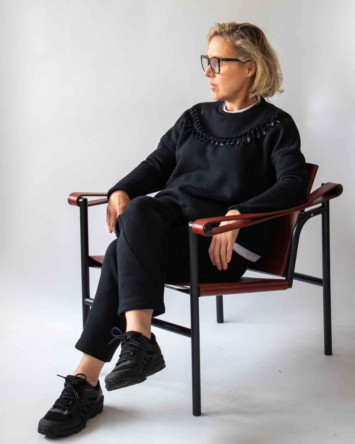 4.	Woman sitting relaxed in a leather and wood designer chair wearing the Asmuss Anni Sweatshirt in Black and the Curve Joggers in Black with a white background
