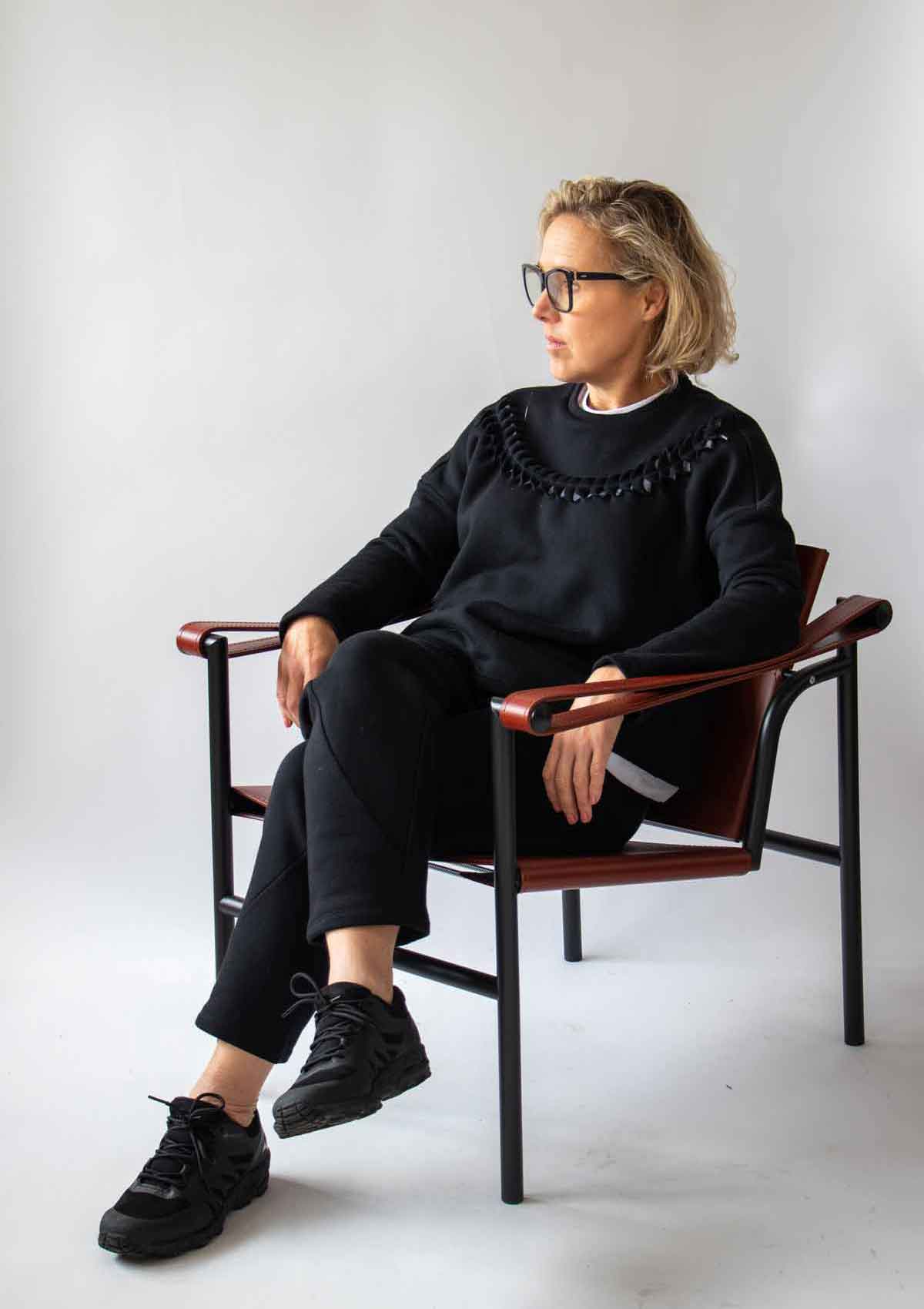 4.	Woman sitting relaxed in a leather and wood designer chair wearing the Asmuss Anni Sweatshirt in Black and the Curve Joggers in Black with a white background