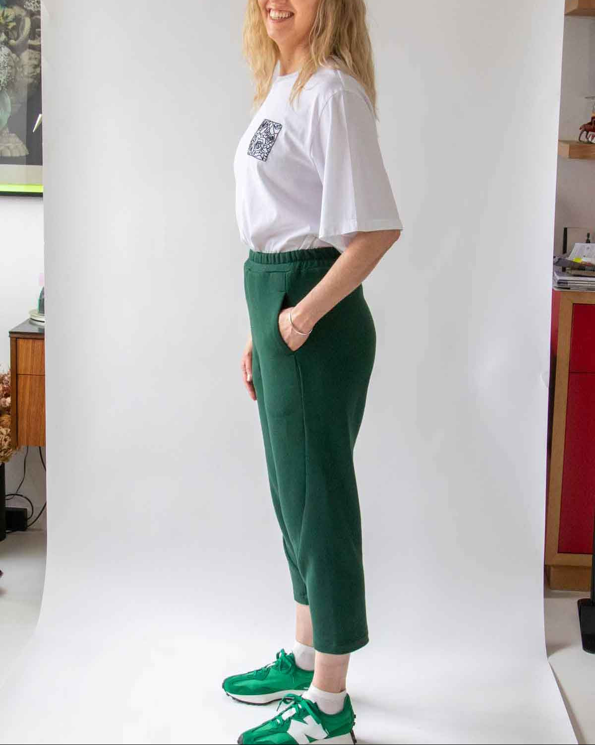 Woman, smiling with long blond hair, standing side on wearing the Asmuss Curve Joggers in Trekking Green and Evelyn Tshirt in White with Black Rose Embroidery and bright green trainers