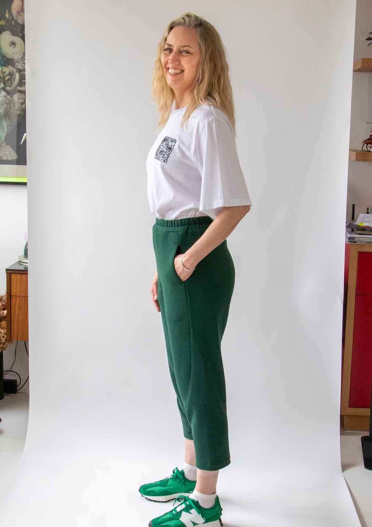 Woman, smiling with long blond hair, standing side on wearing the Asmuss Curve Joggers in Trekking Green and Evelyn Tshirt in White with Black Rose Embroidery and bright green trainers