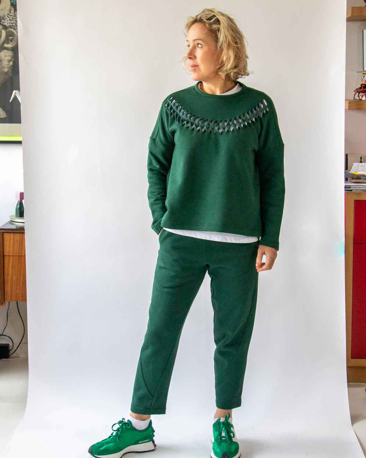 Woman, with short wavy blond hair, wearing the Asmuss Curve Joggers in Trekking Green and the Anni Sweatshirt in Trekking Green, standing with weight on one foot and looking to her right