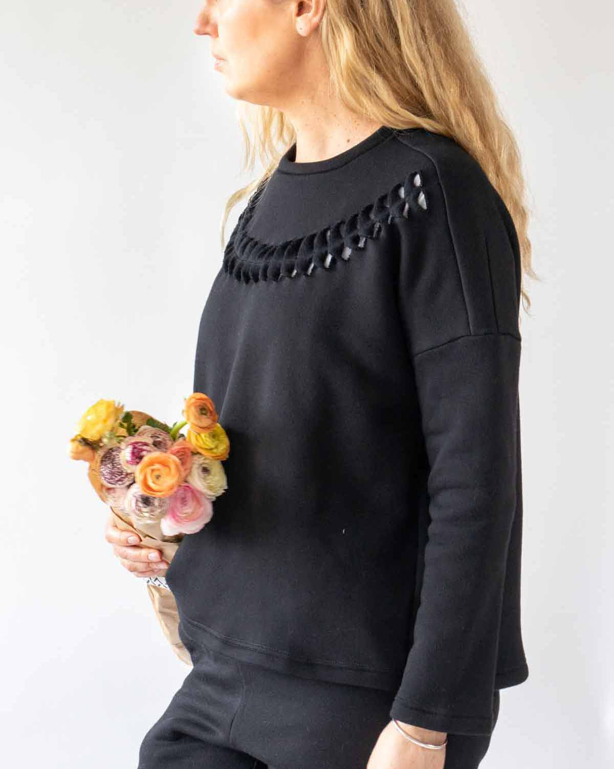 Woman with long wavy blonde hair standing side on wearing the Anni Sweatshirt in Black with cut twist and stick detailing on the front. She is carrying a paper wrapped bunch of flowers in yelllow, pink and orange