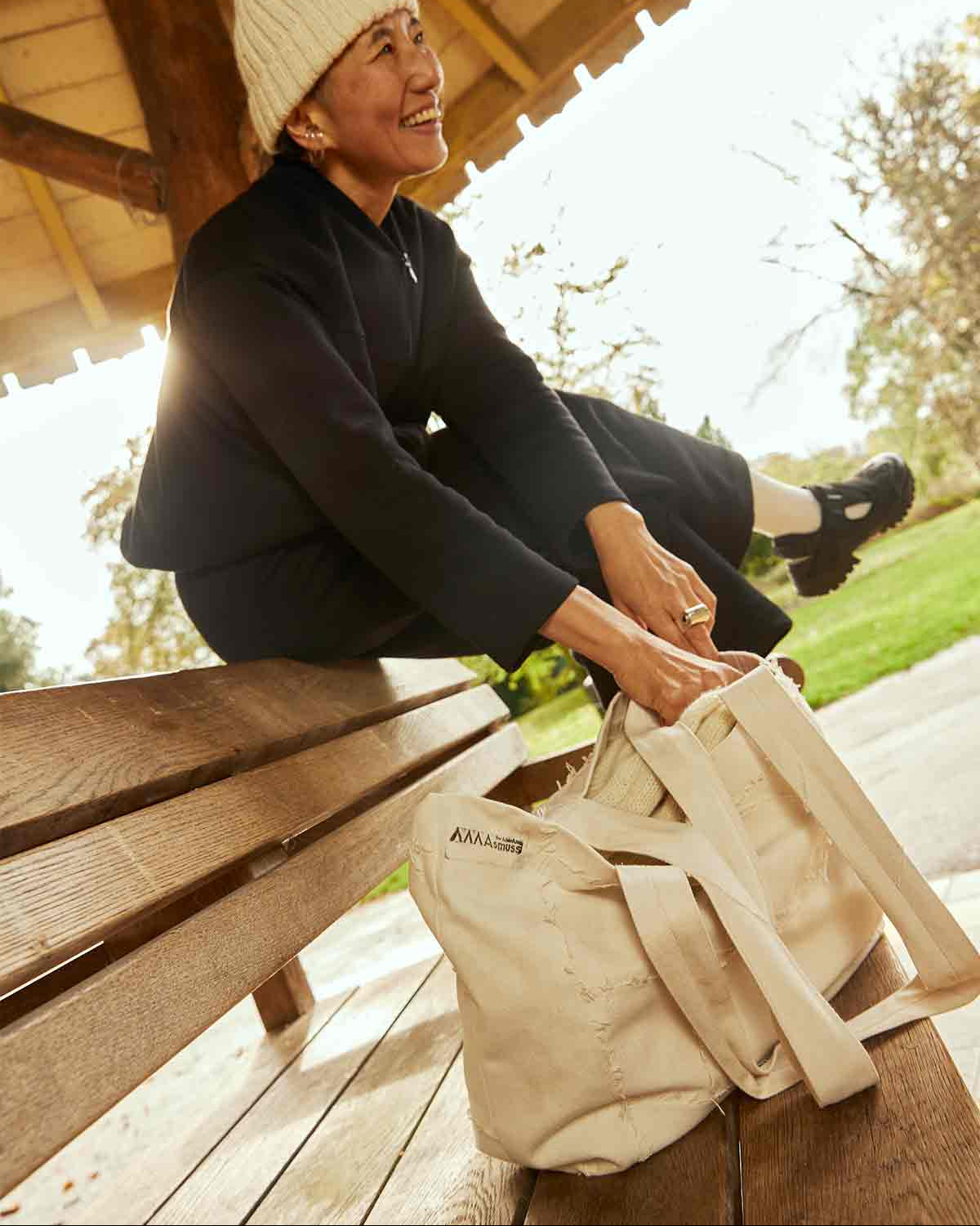 Woman sitting on bench in gazebo, wearing the Asmuss Hepworth zip through sweat, Column joggers in black and also an Asmuss hand knitted ecru beanie. The women is laughing and reaching down into the Asmuss Scraps Tote bag