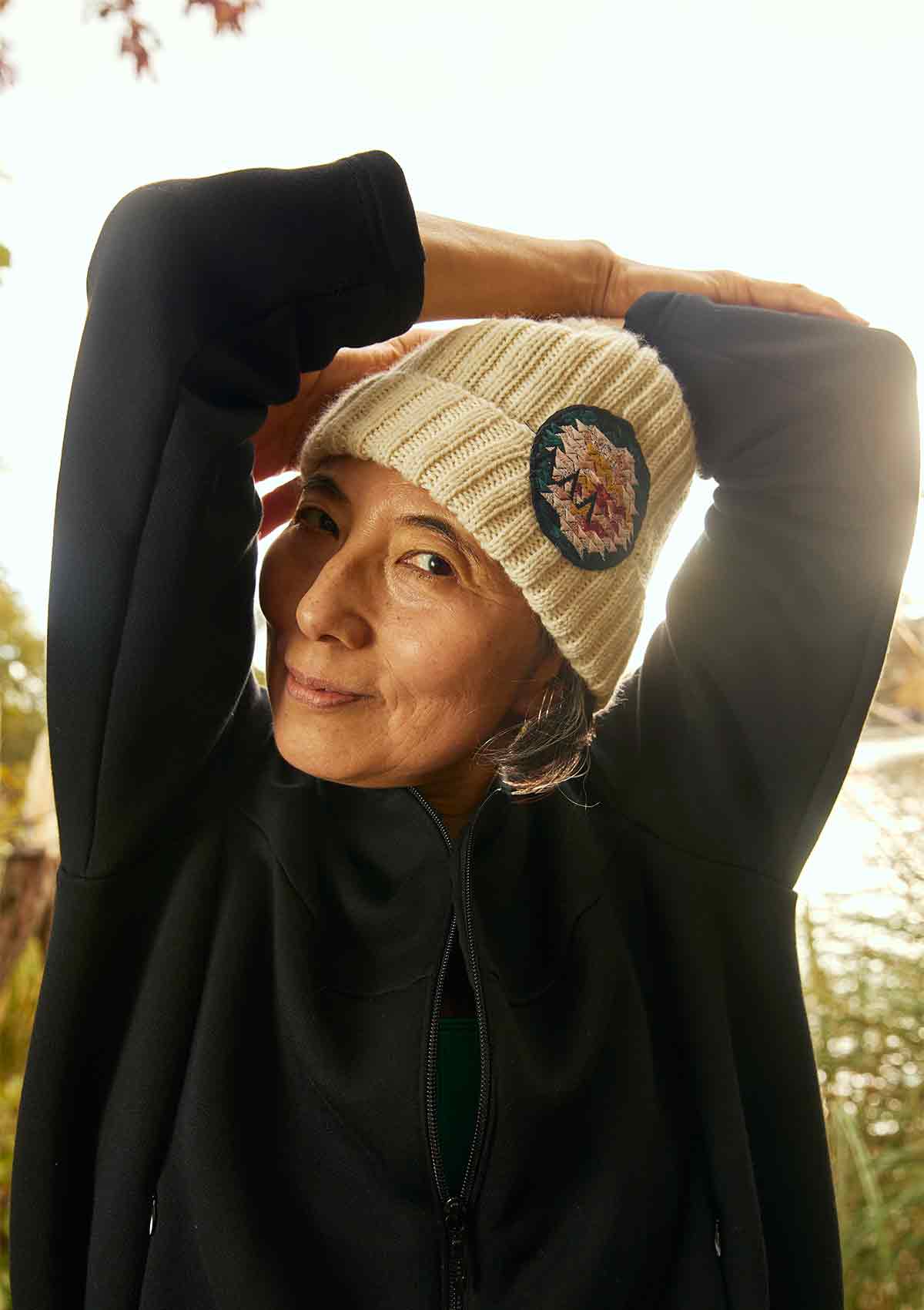 Woman with arms above her head framing the Asmuss Hand Knitted Beanie in Natural White with geometric rose embroidery badge, her head is turned to the left