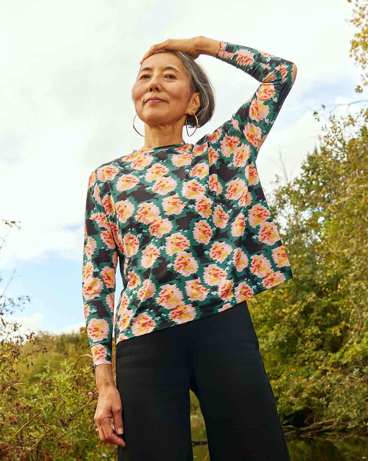 Woman wearing the Asmuss Paige Long Sleeve T-shirt in geometric rose print. She has one hand on her head and is standing in front of trees