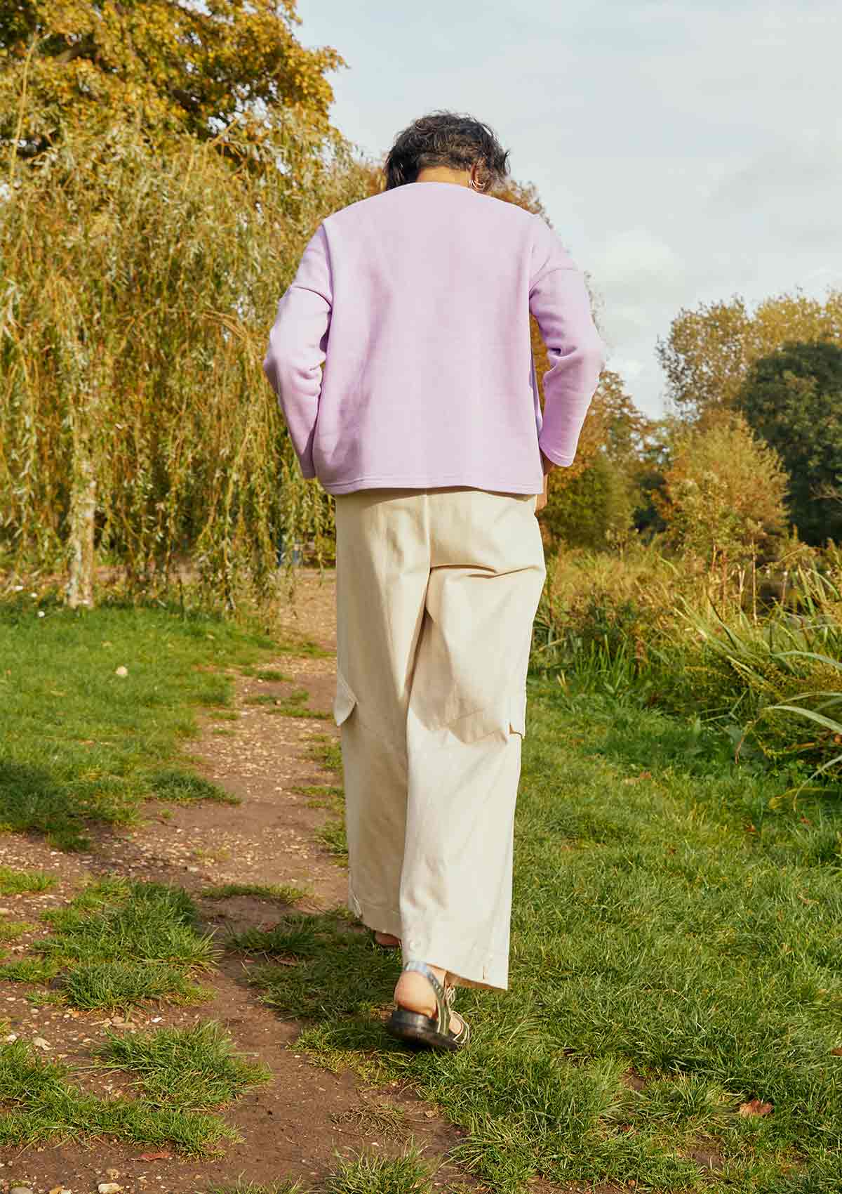Woman walking away through a field with willows. She is wearing the Asmuss Eddy Cargo Trousers in ecru and the Asmuss Anni Sweatshirt in Lilac.