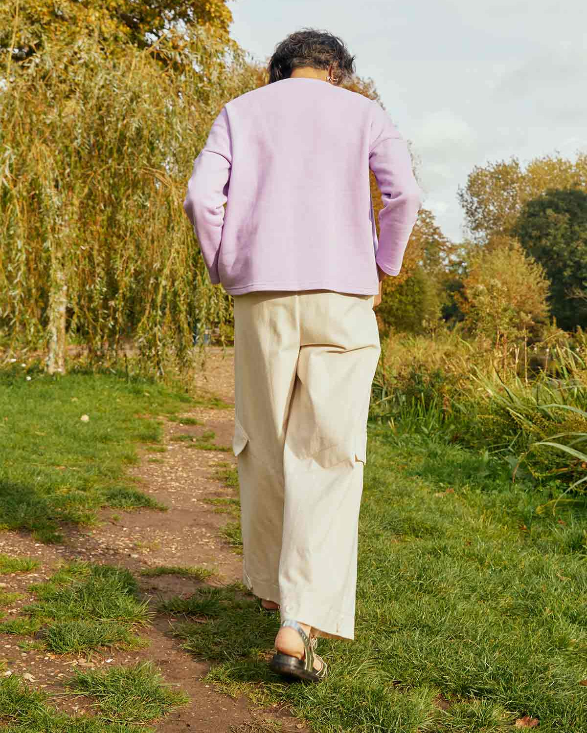 Woman walking away through a field with willows. She is wearing the Asmuss Eddy Cargo Trousers in ecru and the Asmuss Anni Sweatshirt in Lilac.