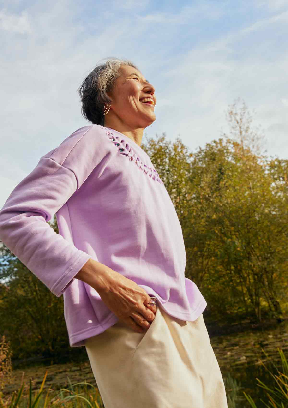 Woman smiling and laughing standing side on wearing the lilac Asmuss Anni Sweatshirt.  She is standing in front of trees and a pond