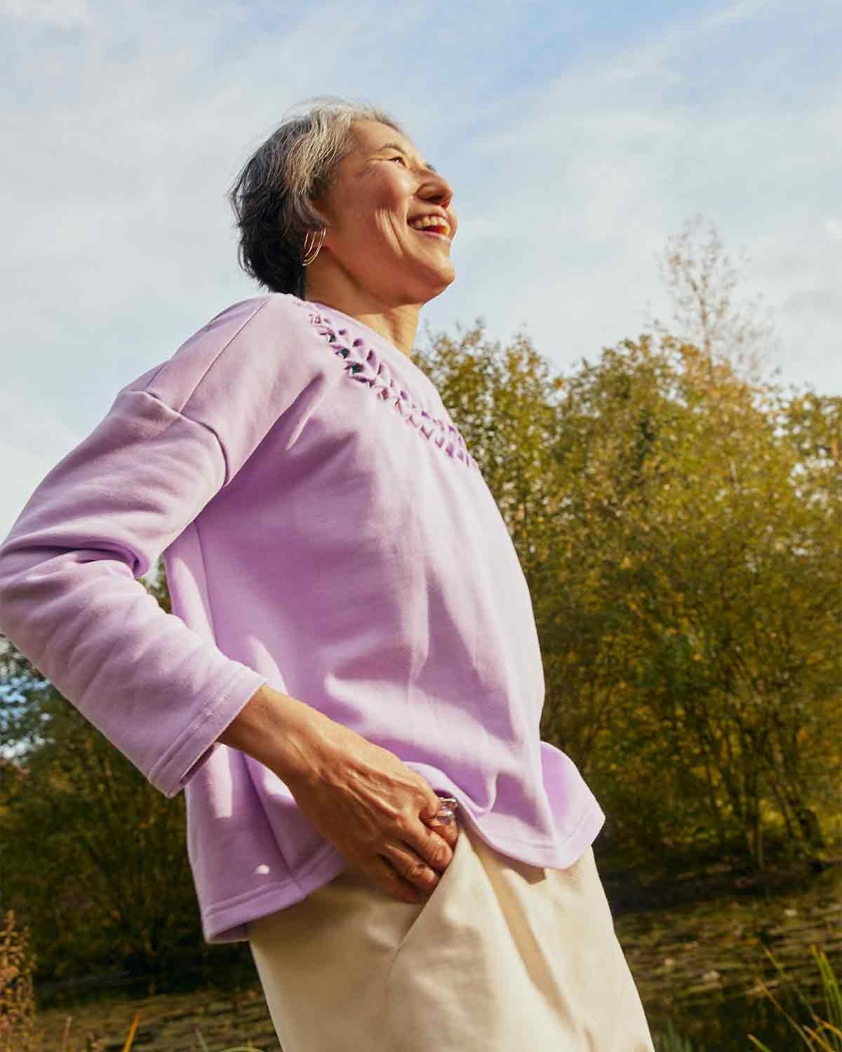 Woman smiling and laughing standing side on wearing the lilac Asmuss Anni Sweatshirt.  She is standing in front of trees and a pond