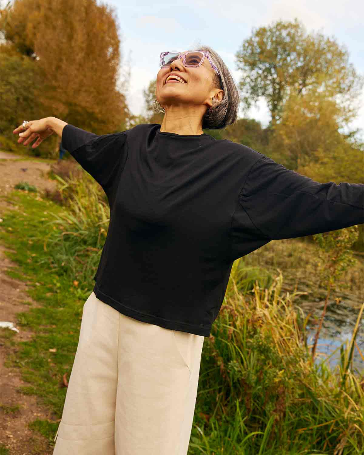 Woman looking up into the sky with her arms out.  You can see the bat wing sleeves of the Asmuss Puzzle Top that she is wearing.  She is standing in a field with trees and pond behind her.
