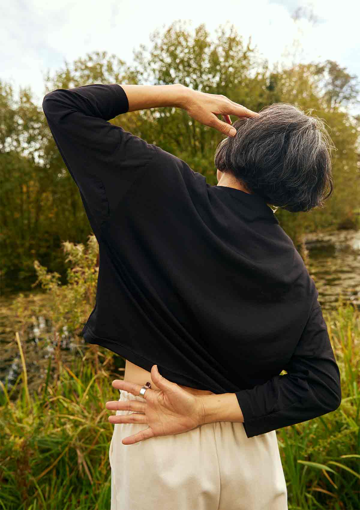 Woman standing facing away with one hand on her head and other bent behind her back.  She is wearing the Asmuss Puzzle Long Sleeve Top in black.  She is stading in front of trees and a pond.