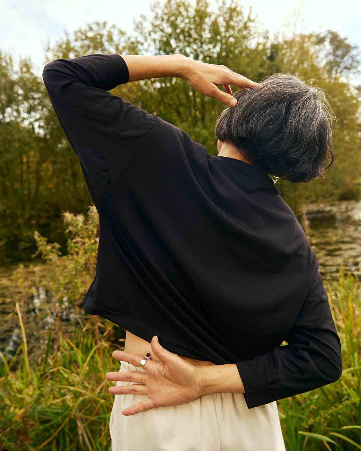 Woman standing facing away with one hand on her head and other bent behind her back.  She is wearing the Asmuss Puzzle Long Sleeve Top in black.  She is stading in front of trees and a pond.
