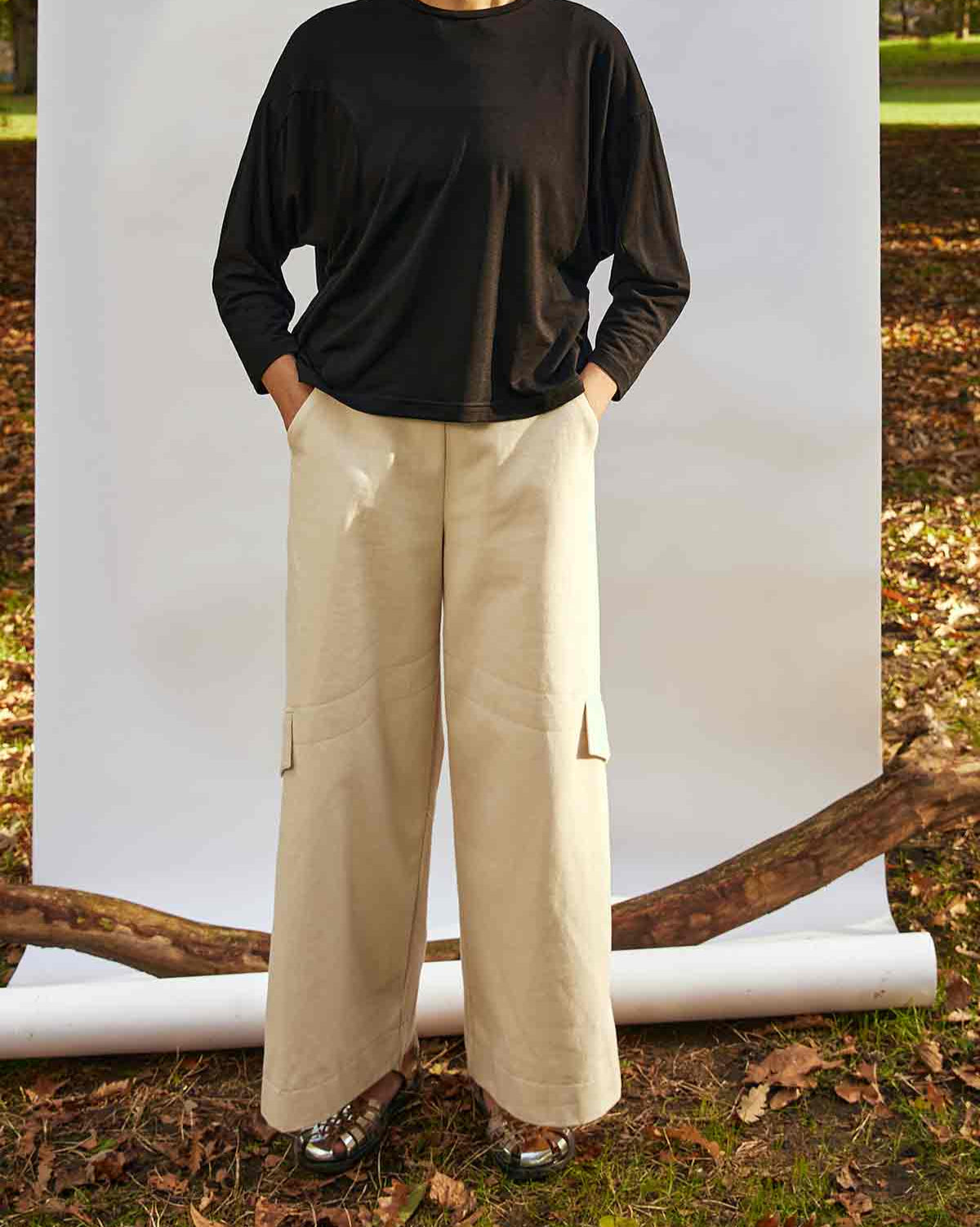 Woman standing face on wearing the Asmuss Eddy Cargo Trousers  and Asmuss Puzzle Top.  The hem buttons are loose.  She is standing in front of a white background, weighted down by a branch in a park
