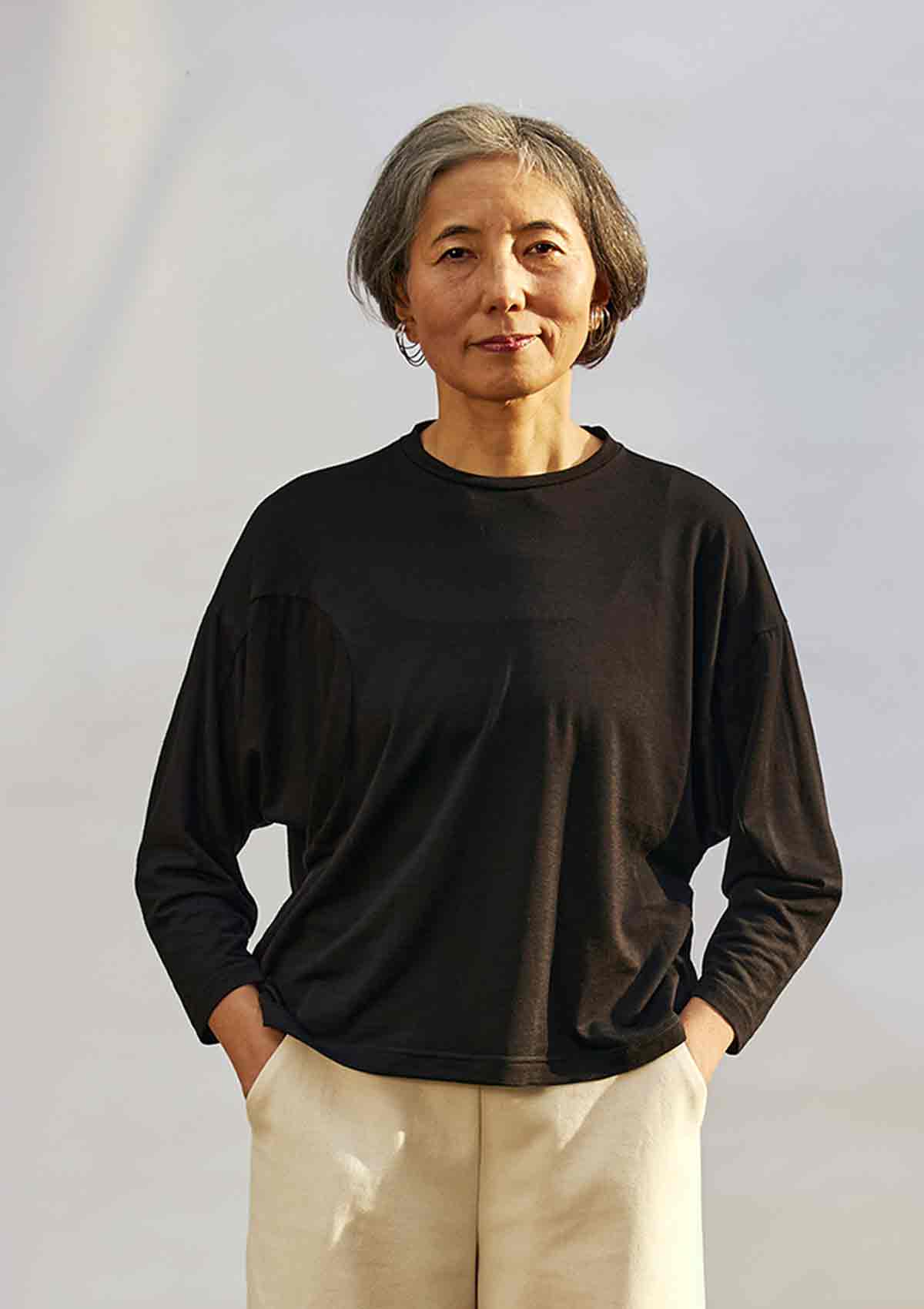 Women staning in front of a white background with the sunning coming fron the right.  She is wearing the Asmuss Puzzle Long Sleeve Top in black.  The sun is highlighting how the fabric falls.