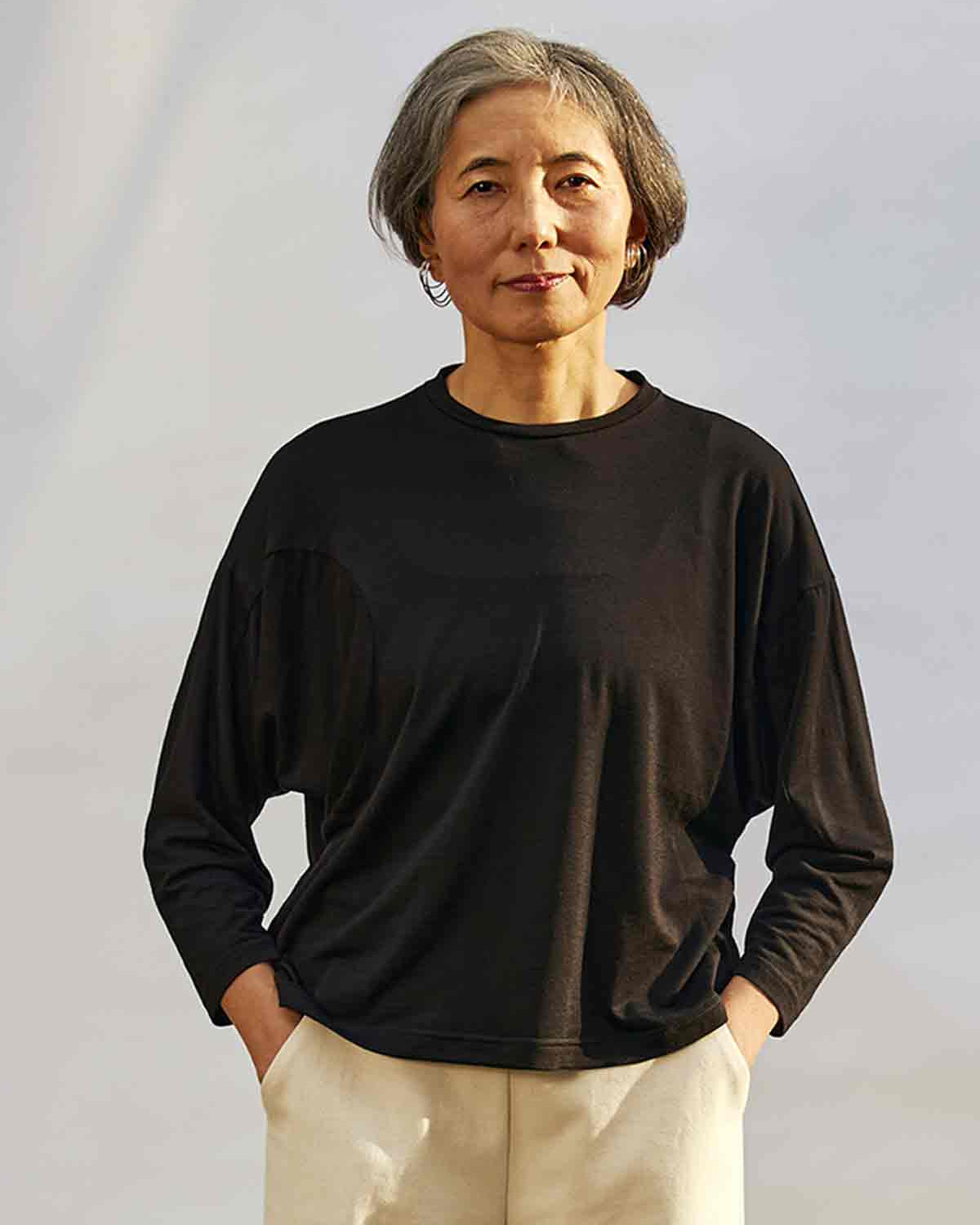 Women staning in front of a white background with the sunning coming fron the right.  She is wearing the Asmuss Puzzle Long Sleeve Top in black.  The sun is highlighting how the fabric falls.