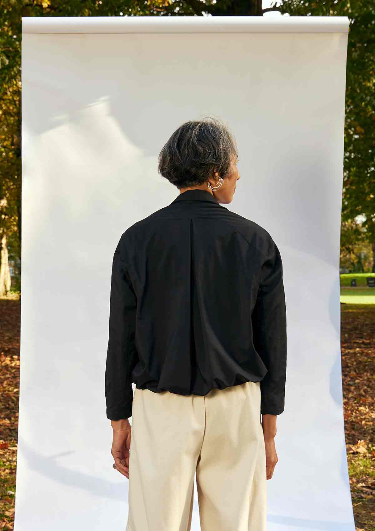 Women, standing back on, wearing the Asmuss Roam Jacket showing the inverted pleat detail on the back, with her hands down by her sides. She is standing in front of a whte background in a park